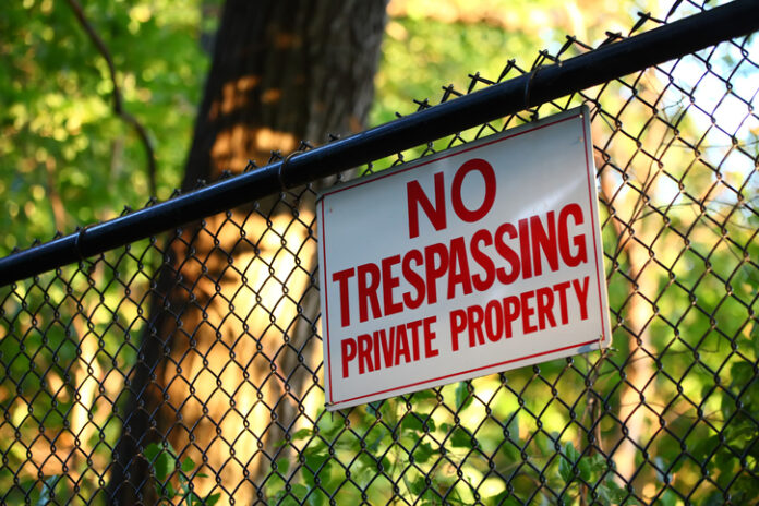 A close-up image of a 'No trespassing - private property' sign hanging on a fence with green trees on the background