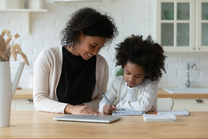 African American young mother and little daughter sit at desk in kitchen studying online together, biracial mom and small girl child handwrite, do homework, learning at home, homeschooling concept