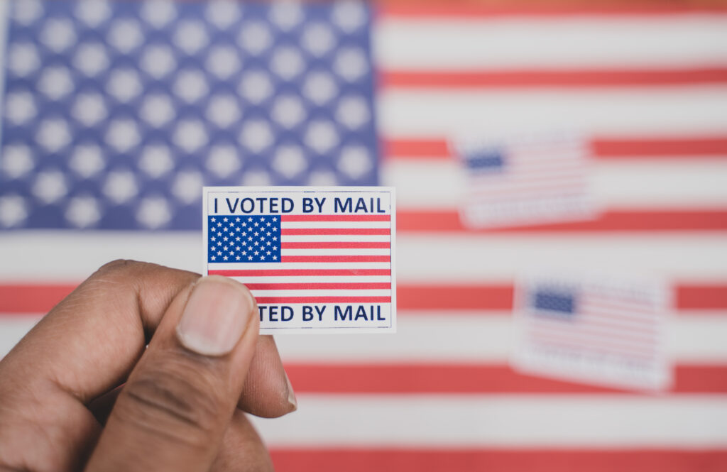 Mail-in Ballot in front of an American flag with an "I voted by mail" sticker