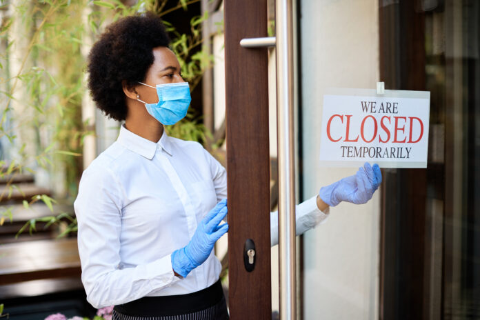 Black owner wearing protective face mask while hanging closed sign at entrance door of her cafe during COVID-19 epidemic.