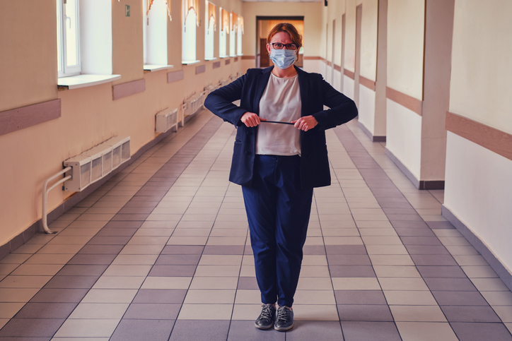 An angry teacher in a medical mask is standing in an empty school corridor. Female teacher during coronavirus quarantine
