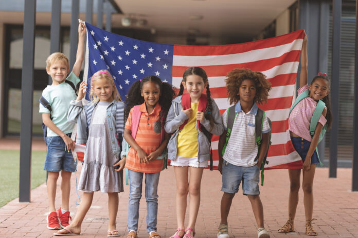 Front view of happy school students standing in outside corridor at school while holding american flag