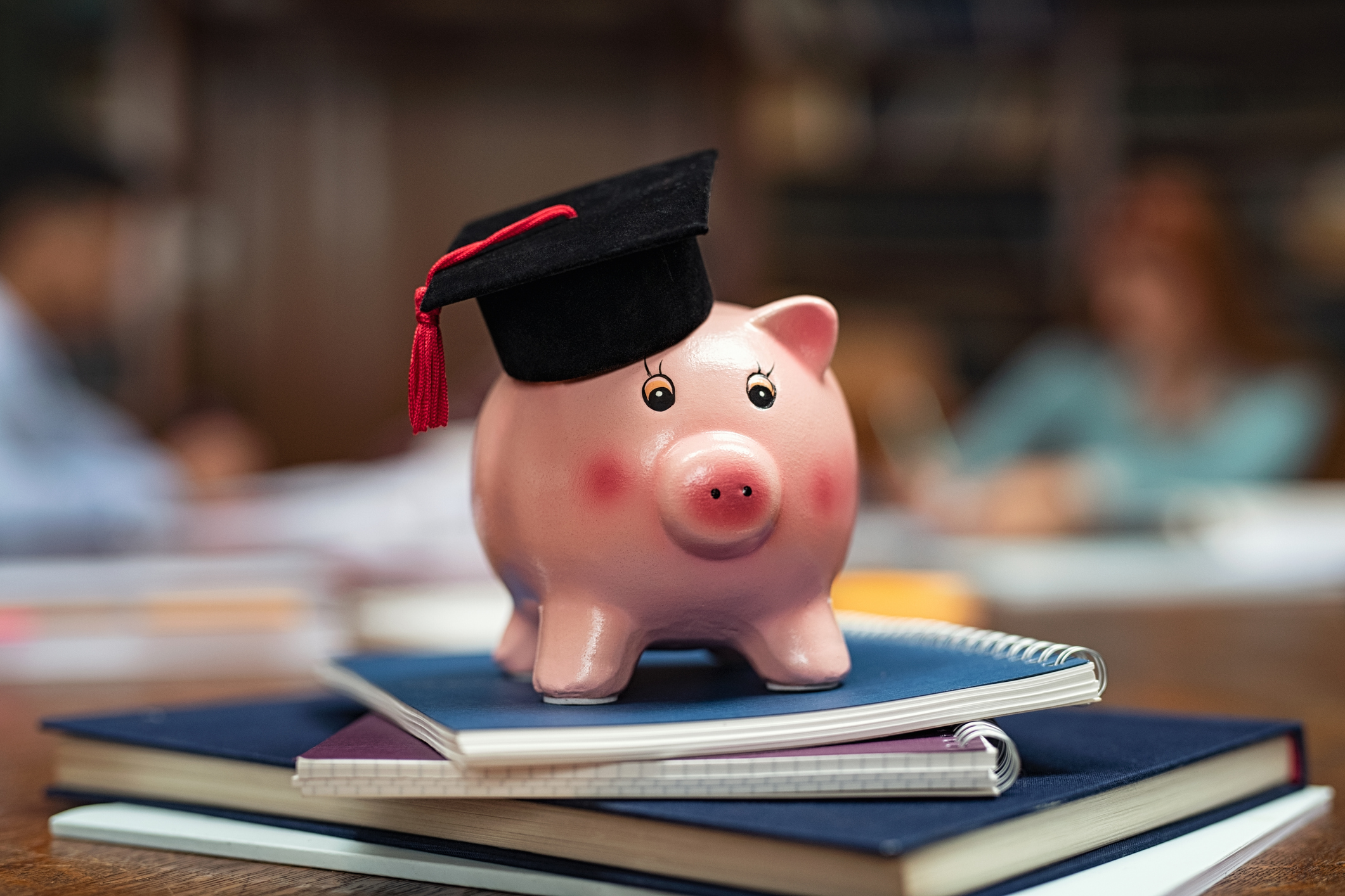 Piggy bank wearing mortar board on pile of books in library while students studying in background. Saving money for college with piggy bank on books in library. Savings and education debt concept. Student loan