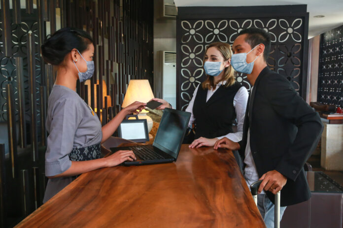 Couple and receptionist at counter in hotel wearing medical masks as precaution against virus. Young couple on a business trip doing check-in at the hotel, hospitality industry