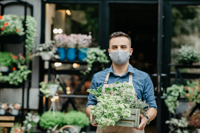 Small business and start of working day. Man in protective mask takes out box of plants outside in front of flower shop