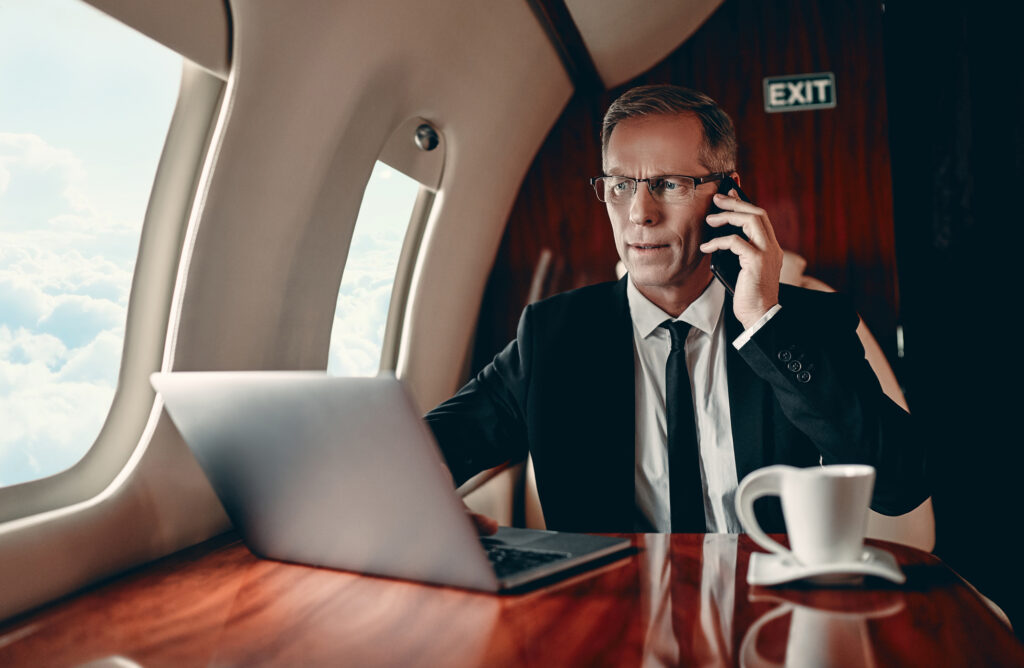 Rich businessman in suit is working on a laptop and speaking on mobile phone while flying in private jet.