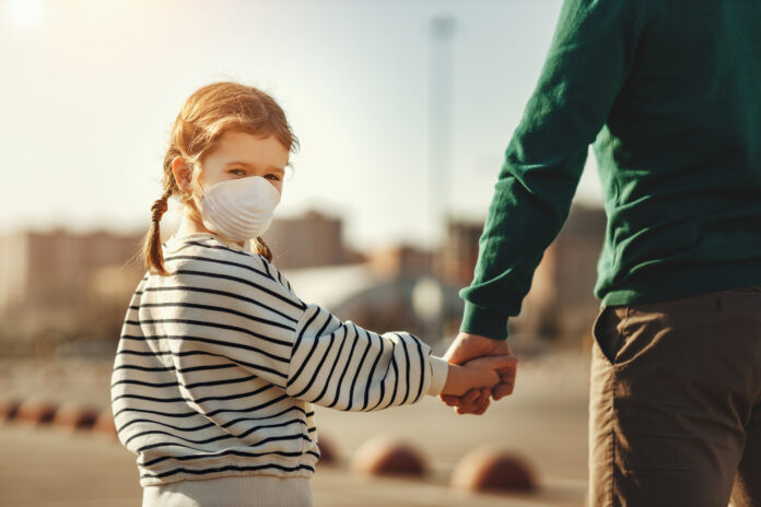 Back view of little girl in medical mask looking at camera over shoulder and holding hand of crop father while walking on parking lot during coronavirus outbreak, parenting