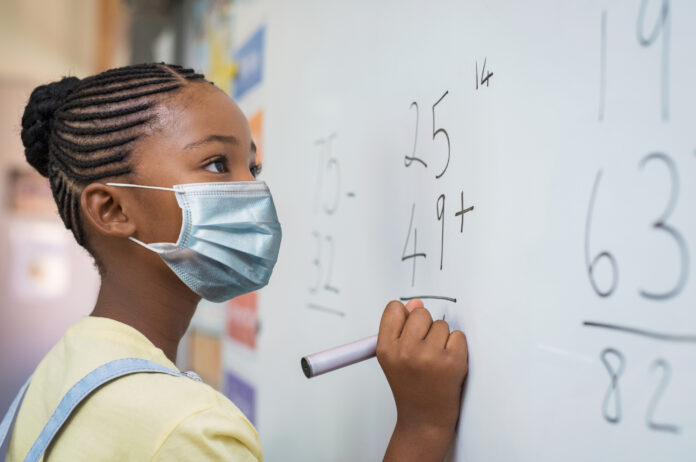 Portrait of african girl wearing face mask and writing solution of sums on white board at school. Black schoolgirl solving addition sum on white board during Covid-19 pandemic. School child thinking while doing mathematics problem and wearing surgical mask due to coronavirus.