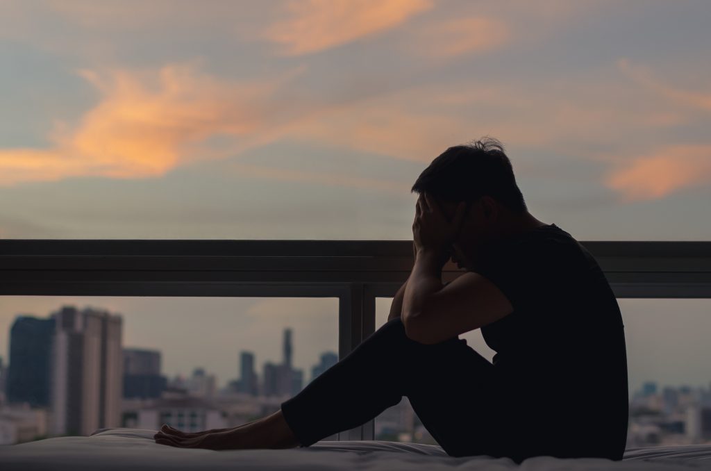 A person sitting and feeling depressed on bed with city view in dusk moment. Stay home, depression and loneliness and mental health concept.