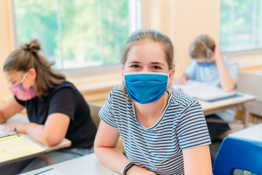 A thirteen year old girl is sitting at her desk in class with others students around her. She is wearing a mask to protect herself from Covid-19. She is looking at the camera. The others students around her are working.
