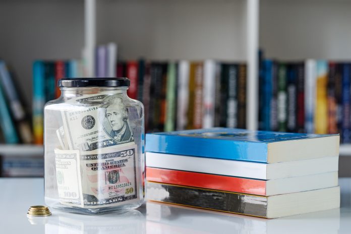 Glass jar full of american us dollars banknotes money and books on the white table in front of the shelf savings and study scholarship concept wealth as knowledge investment