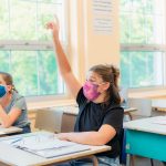 Back-to-School Mask Mandates Are Outraging Parents—Here’s How to Get Around Them A thirteen year old girl is sitting at her desk in class with others students around her. She is wearing a mask to protect herself from Covid-19. She is looking at the camera. The others students around her are working.