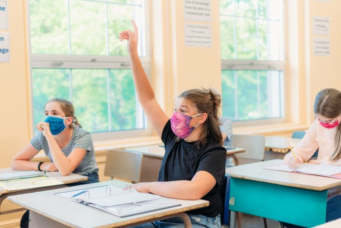 A thirteen year old girl is sitting at her desk in class with others students around her. She is wearing a mask to protect herself from Covid-19. She is looking at the camera. The others students around her are working.