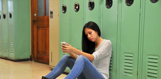 Sad teenager girl sitting against locker in hallway with her cell phone.