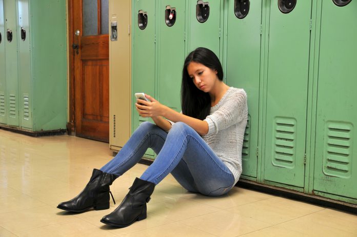 Sad teenager girl sitting against locker in hallway with her cell phone.
