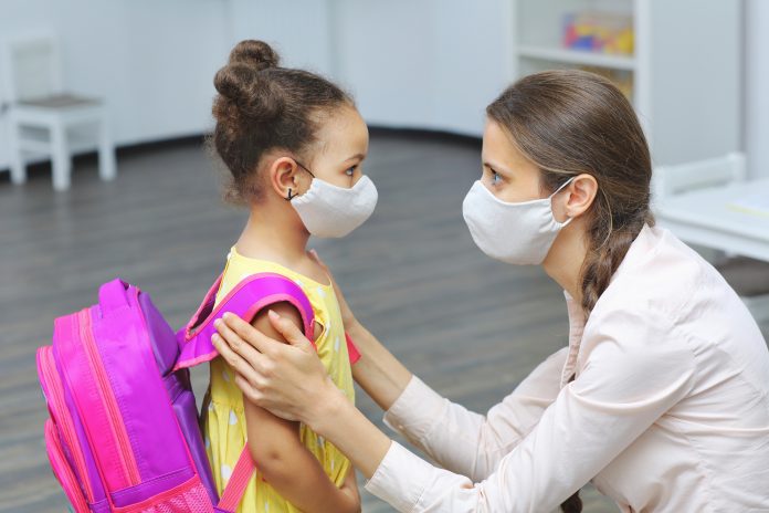 a light skinned female teacher in a medical mask for respiratory protection is talking to a dark skinned student with a mask on her face and a backpack on her shoulders