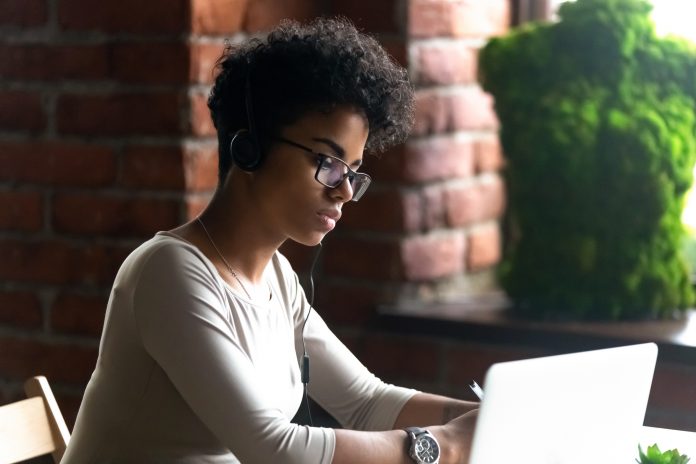 Focused African American woman wearing headphones using laptop, making writing notes, serious female student learning language, watching online webinar, listening audio course, e-learning, education