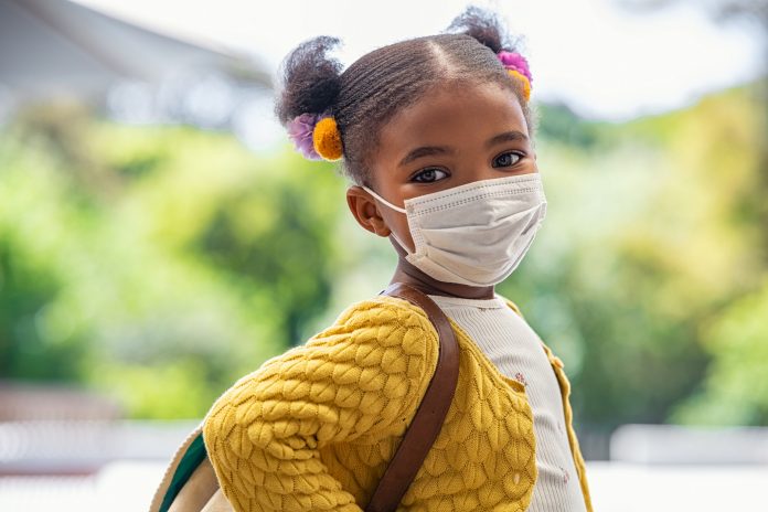 Smiling cute little girl with school backpack and protective face mask ready for first day of school during covid pandemic. African american female child wearing surgical mask while looking at camera. Cute black kid going back to school during coronavirus pandemic disease.