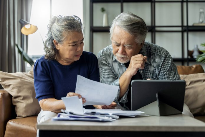 old retired asian senior couple checking and calculate financial billing together on sofa involved in financial paperwork, paying taxes online using e-banking laptop at living room home background