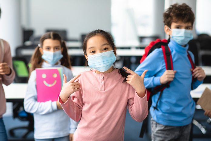 Back To School. Small asian girl pointing at facemask looking at camera standing in classroom with diverse classmates, showing new normal covid quarantine rules due to coronavirus pandemic outbreak